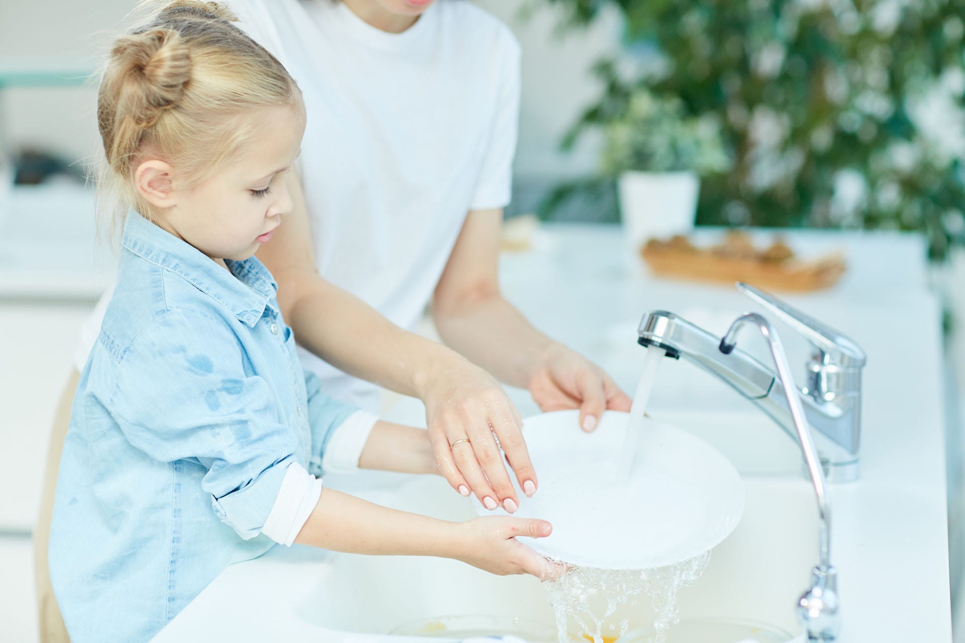 Little girl helping her mother wash dishes over sink in the kitc